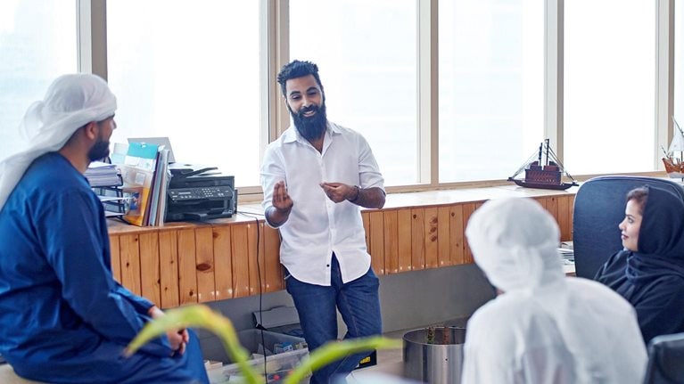 A young businessman in jeans and a button down sharing ideas with male and female colleagues in traditional Emirati clothing. The team is casually seated in a circle in a open office setting.