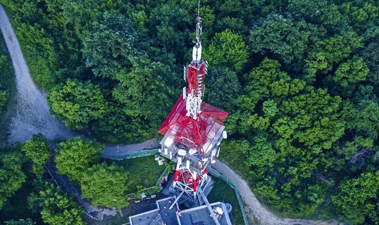 From above, a red telecommunications tower can be seen amidst a lush green forest.