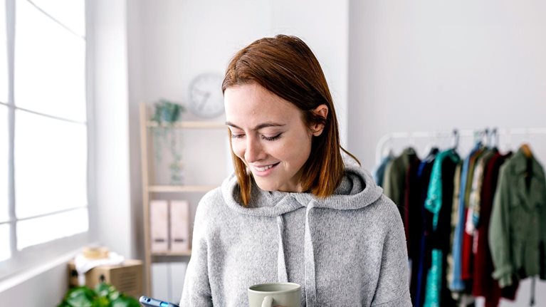 Woman dressed in gray hoodie looking down and phone in one hand, other hand holding ceramic coffee mug.