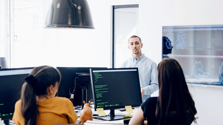 Software development team having a discussion during a presentation in a modern office around computer monitors showing code.