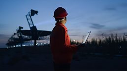 Photograph of a person wearing a red safety helmet and jacket holding a laptop, standing outdoors at dusk near large telecommunications equipment. The scene highlights a work environment with artificial lighting and a darkening sky, suggesting monitoring or inspection activities during low light conditions.