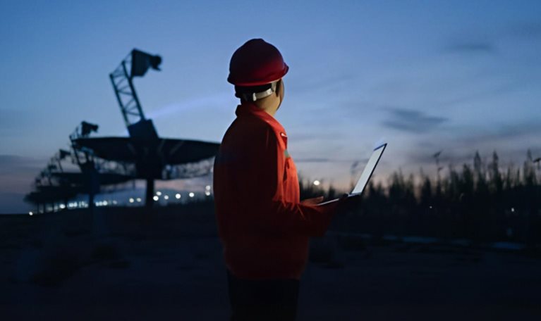 Photograph of a person wearing a red safety helmet and jacket holding a laptop, standing outdoors at dusk near large telecommunications equipment. The scene highlights a work environment with artificial lighting and a darkening sky, suggesting monitoring or inspection activities during low light conditions.