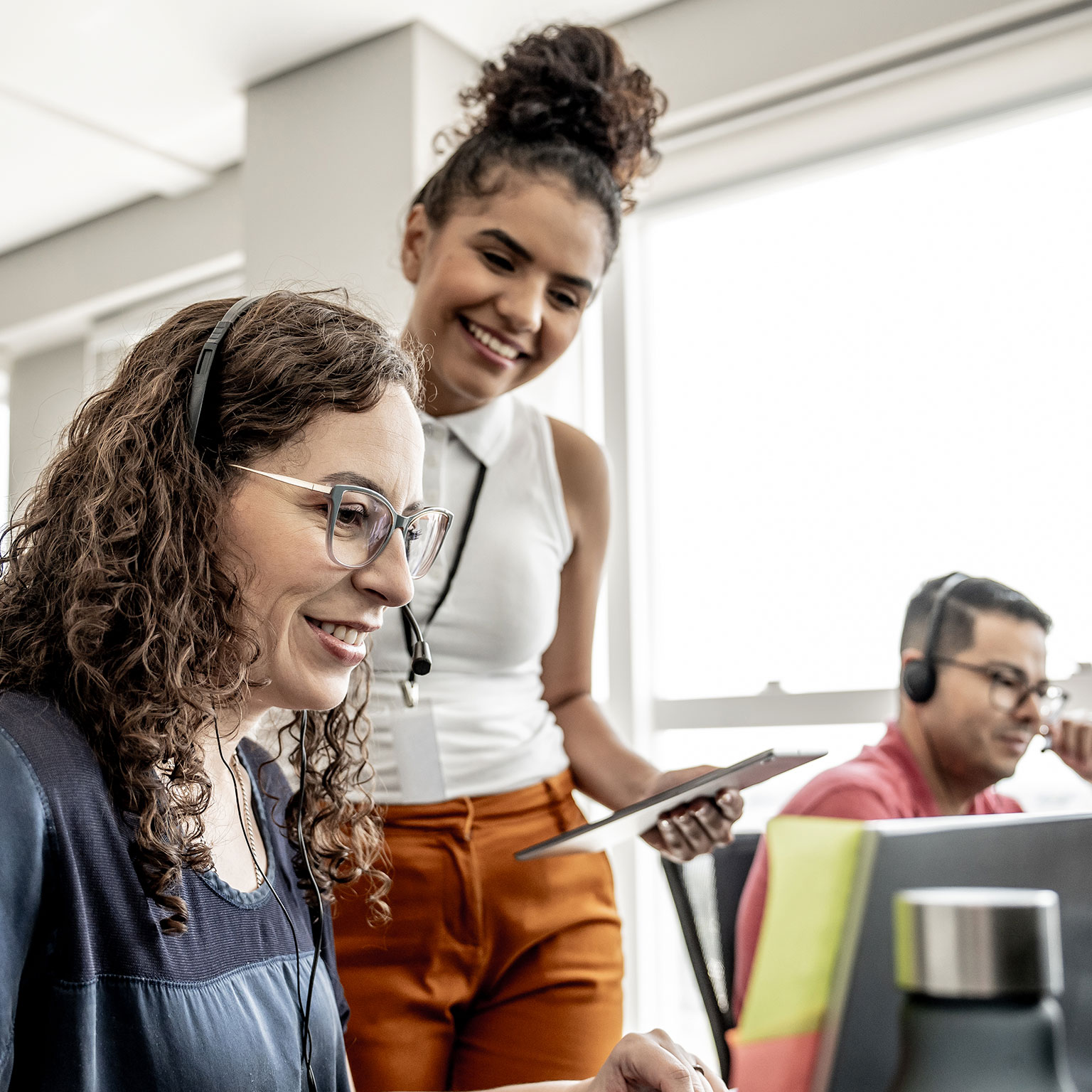 Photograph of a modern office setting with three people engaged in work activities. One person with curly hair sits at a desk, another stands holding a tablet, and a third works on a laptop, highlighting a collaborative and technology-driven work environment.