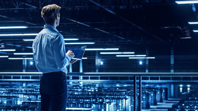 A person standing on an elevated walkway in a large, blue-lit data center, holding a laptop and overlooking rows of server racks.
