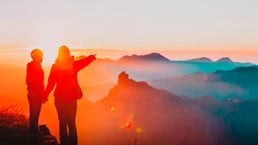 mother holding hand of son while pointing over canyon rim, sun setting in background - photo
