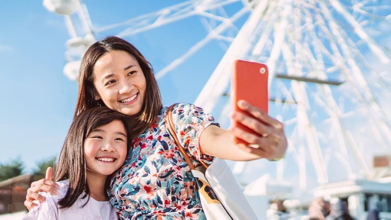 Mom and daughter taking selfie against a ferris wheel when enjoying spending time together in theme park - stock photo