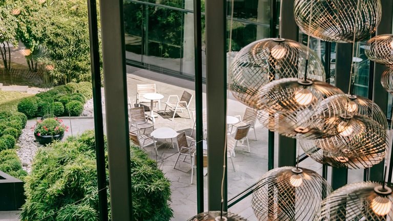 View from inside an Accor hotel through a modern glass wall and golden light fixtures out onto patio seating surrounded by greenery.