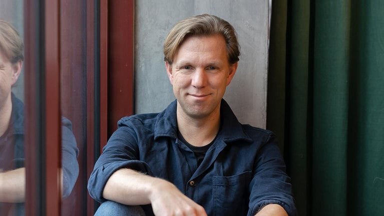 Portrait of Pelle Sommansson in jeans and a collared shirt. Sommansson has a relaxed pose and is leaning against an office window.