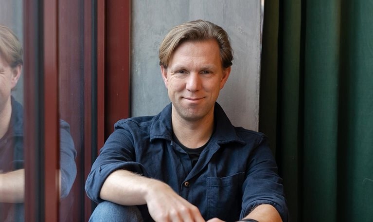 Portrait of Pelle Sommansson in jeans and a collared shirt. Sommansson has a relaxed pose and is leaning against an office window.