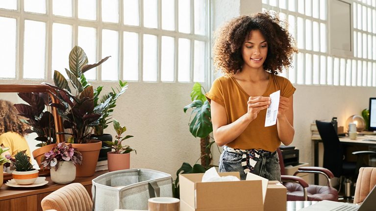 Confident businesswoman holding sticky bill. Young female entrepreneur is working at home office. Packages are on table.
