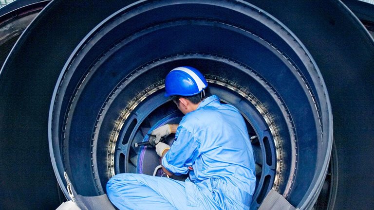 A maintenance technician in protective gear works inside a large jet engine in an aircraft hangar.