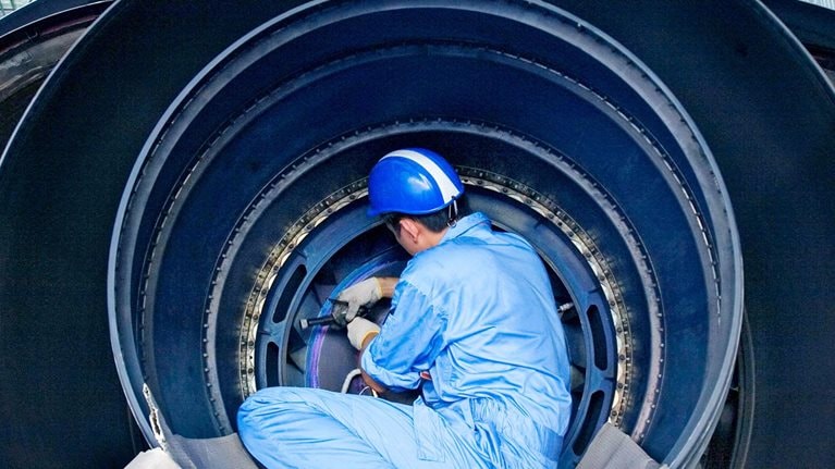 A maintenance technician in protective gear works inside a large jet engine in an aircraft hangar.