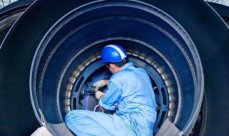 A maintenance technician in protective gear works inside a large jet engine in an aircraft hangar.