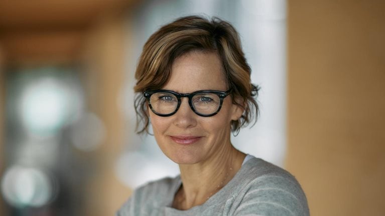 Jennifer Pahlka, donning a light smile and glasses, clad in a gray sweater with white stripes, stands with her arms folded in front of a blurred office setting.