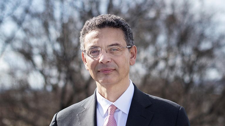 Pedro Conceição, in a suit and tie, standing outdoors in front of a blurred background of winter trees.