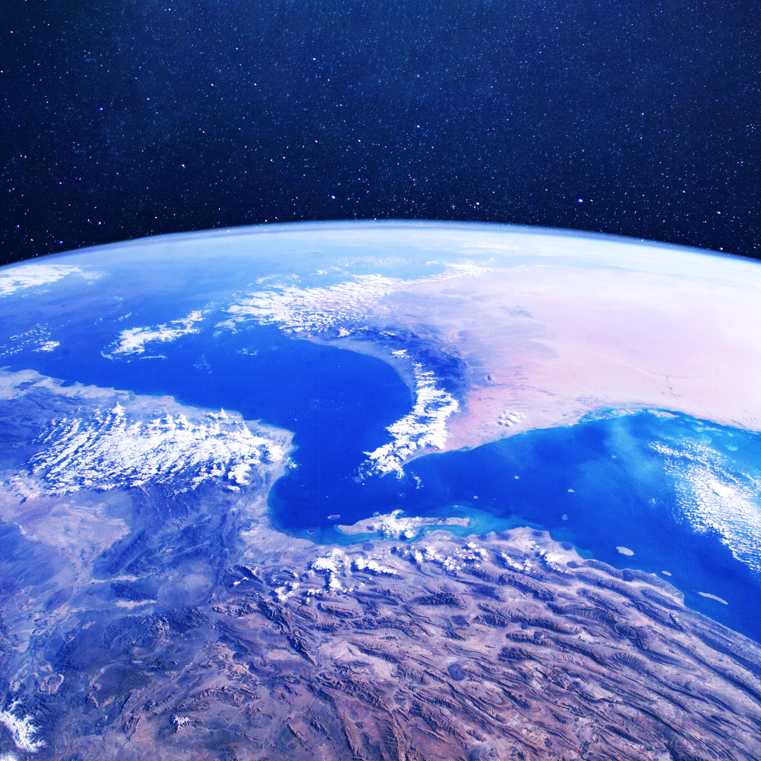 A view of Earth from space centered on the Strait of Hormuz. The curve of the planet, deep blue seas, desert landmasses, and scattered clouds are visible.  