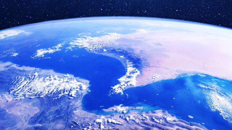 A view of Earth from space centered on the Strait of Hormuz. The curve of the planet, deep blue seas, desert landmasses, and scattered clouds are visible.  