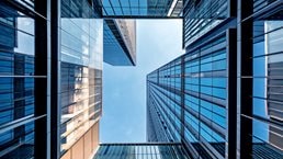 A dramatic upward view between modern glass skyscrapers, their reflective blue surfaces forming a geometric frame around the sky.