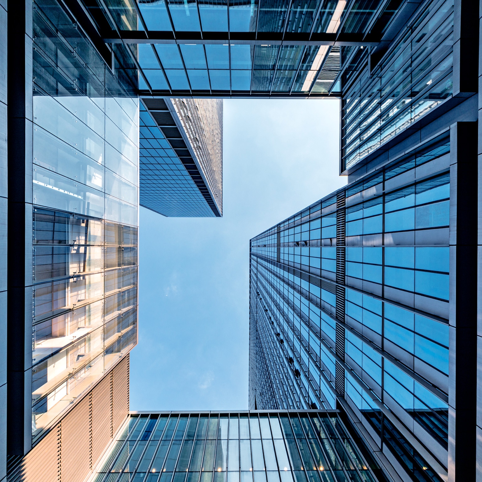 A dramatic upward view between modern glass skyscrapers, their reflective blue surfaces forming a geometric frame around the sky. 