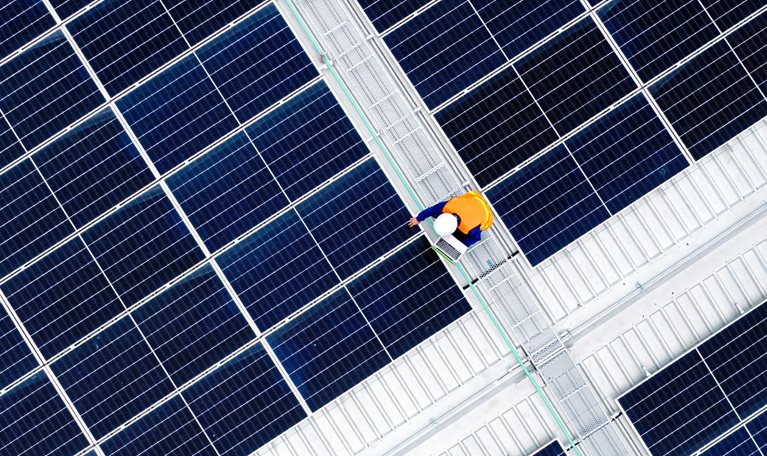 A vast array of dark blue solar panels arranged in a precise geometric grid, viewed from above. At the center, a worker in a bright orange safety vest walks along a maintenance pathway.