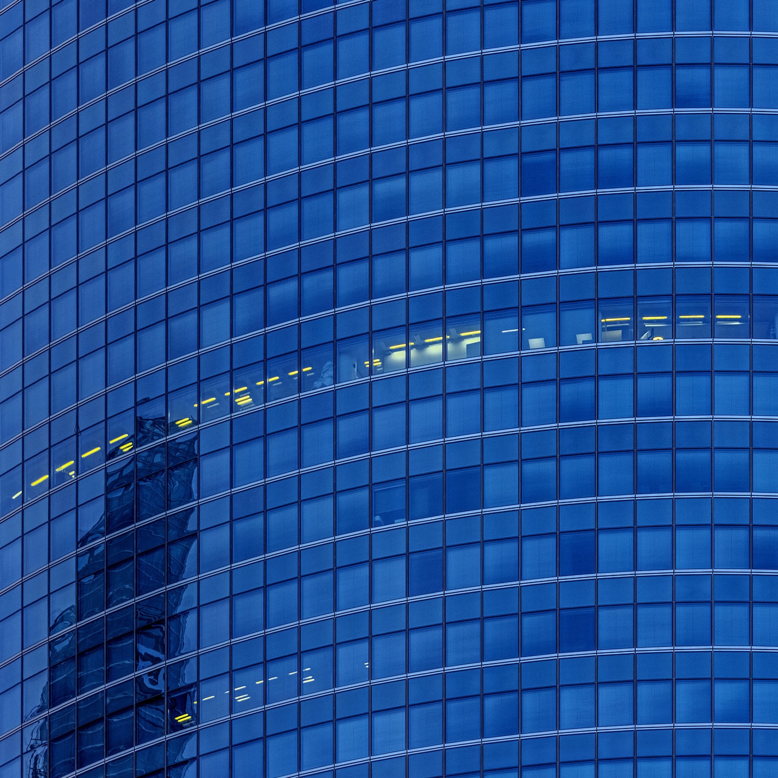 A detail shot of a glass façade skyscraper. All the floors appear dark except for a smattering of offices which have their lights on.
