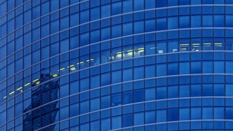 A detail shot of a glass façade skyscraper. All the floors appear dark except for a smattering of offices which have their lights on.