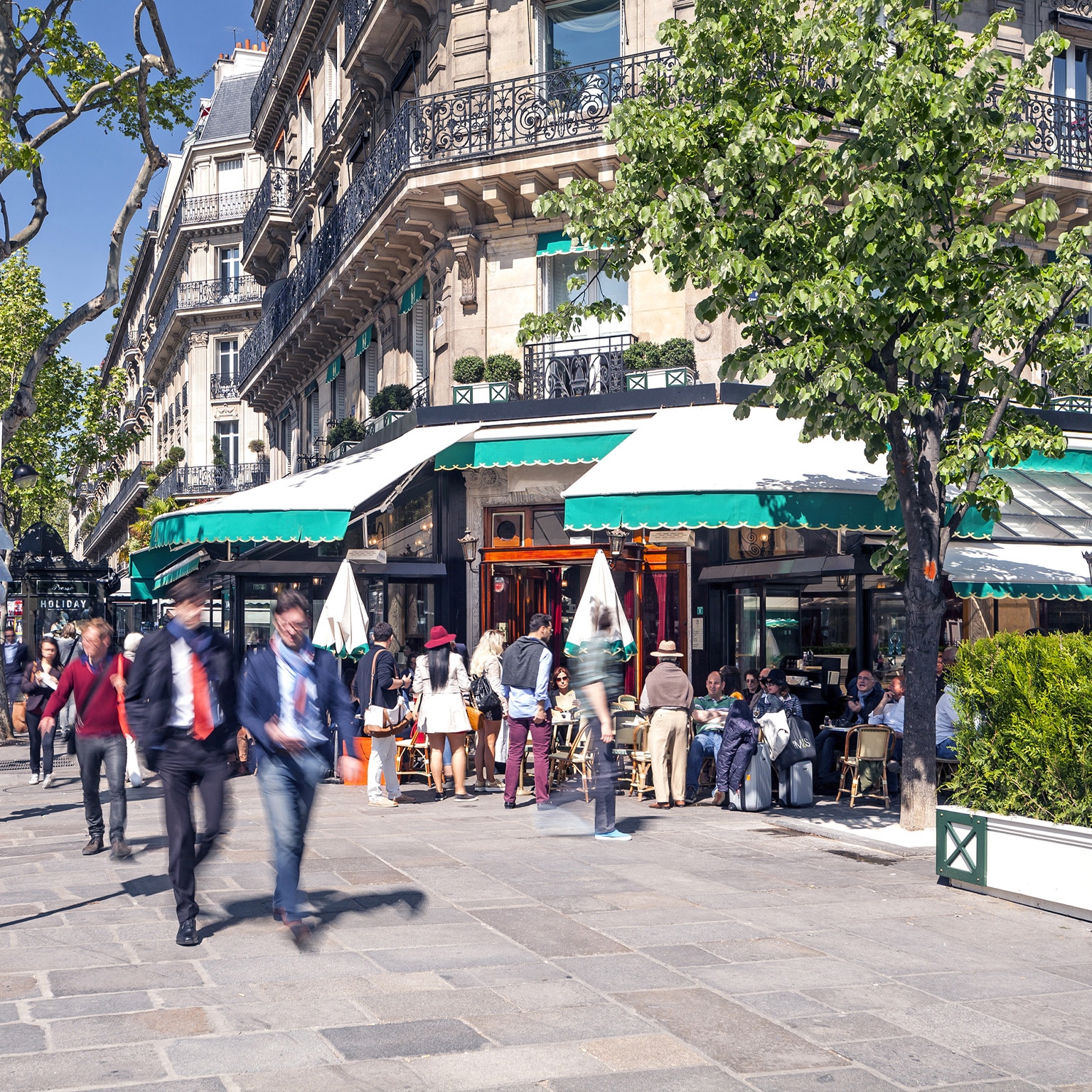 A busy Boulevard Saint-Germain in Paris. A corner café and sidewalk are full of professionals and people going about their day. Apartments are seen above the lower shops.