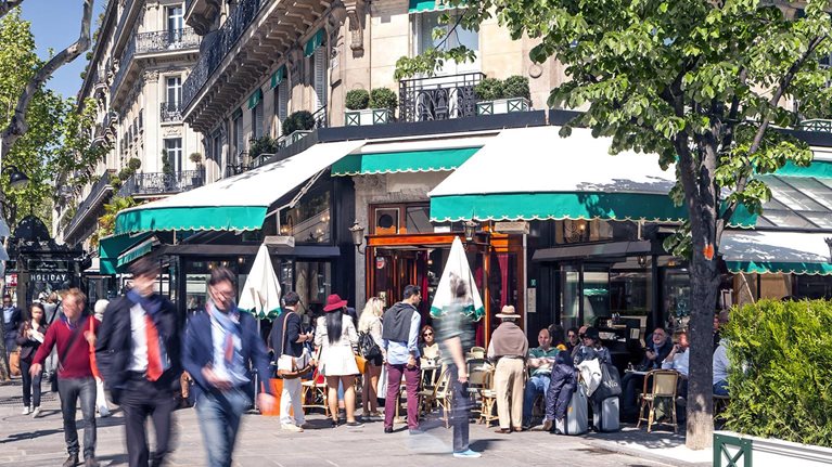 A busy Boulevard Saint-Germain in Paris. A corner café and sidewalk are full of professionals and people going about their day. Apartments are seen above the lower shops.