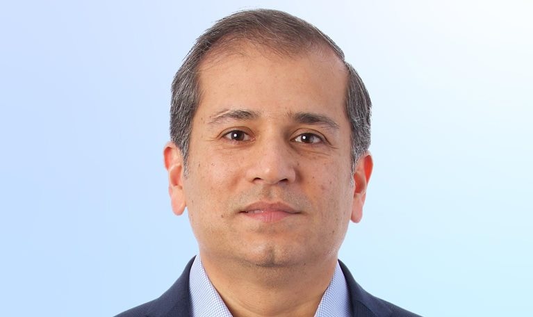 A head-and-shoulders portrait of a Shubham Singhal facing forward with a neutral expression. He is wearing a dark blazer over a light blue patterned dress shirt, set against a soft gradient background that transitions from light gray to pale blue.
