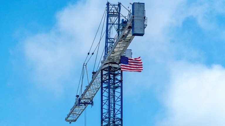 An American flag attached near the top of a tall construction crane waves in the wind. The crane rises against a cloudy blue sky, with its long arm extending outward.