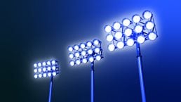 Against a dark blue backdrop, three sets of stadium lights emit a radiant glow, evoking a feeling of excitement and suspense.