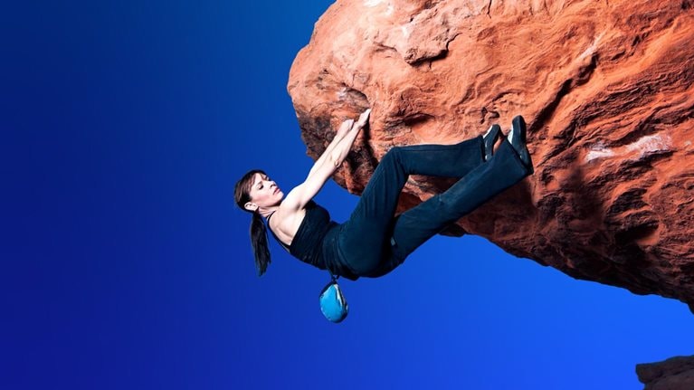 A woman in black rock-climbing clothing navigates a large overhanging bolder without a rope. She has a firm hold with both hands but seeks a foot hold as she makes her way over the protruding rock.