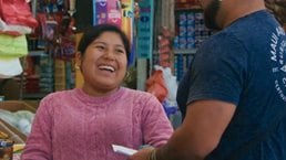Photograph of a small grocery store checkout area showing a cashier wearing a pink sweater and gloves interacting with a customer in a blue shirt and cap. Shelves behind the cashier are stocked with various packaged food items, including biscuits, cereals, and snacks, highlighting a typical local market setting.