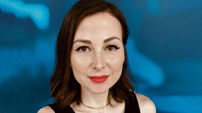 Portrait of Zeinab Hijazi with short dark-brown hair and red lipstick. She is wearing a black tank top and a gold necklace. The background is a blue and white abstract pattern.