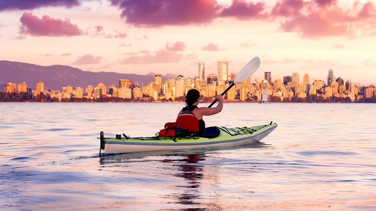 As the sun sets behind the Vancouver skyline, a woman paddles her kayak on a vast body of water. The vibrant sunset sky paints the horizon with shades of pink and purple, casting a mirror-like reflection on the water.