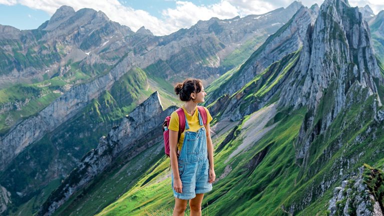 A woman wearing hiking clothes and a red backpack stands on a grassy mountain ridge surrounded by the steep rocky peaks and green valleys of the Swiss Alps.