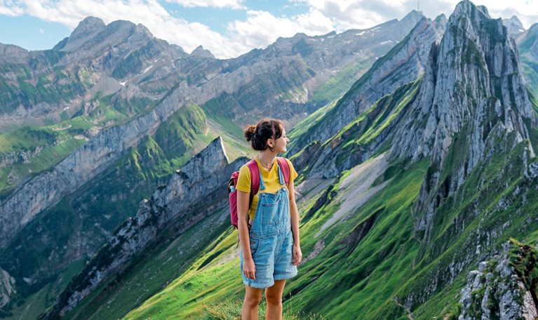 A woman wearing hiking clothes and a red backpack stands on a grassy mountain ridge surrounded by the steep rocky peaks and green valleys of the Swiss Alps.