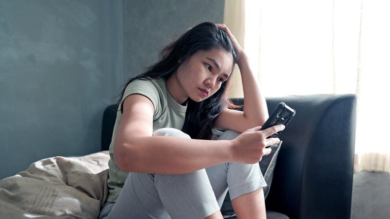 A teenager girl sitting on a day bed and looking at a phone with a concerned expression. 
