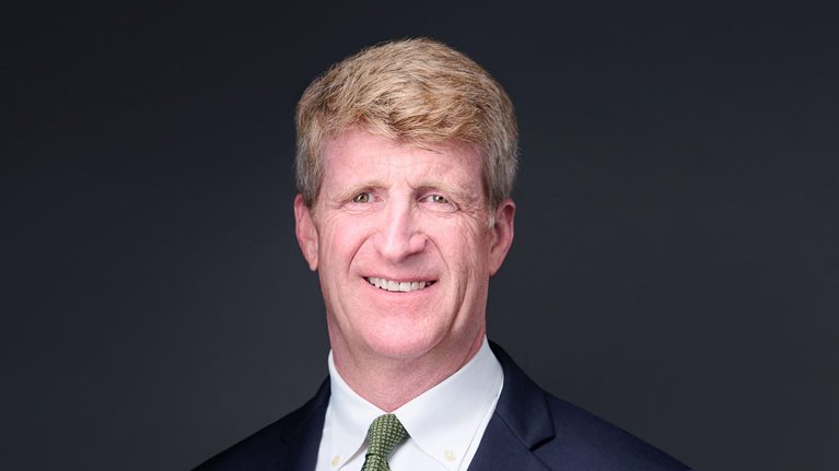 Patrick J. Kennedy, wearing a navy suit jacket, a green tie, and a smile, photographed against a dark background.