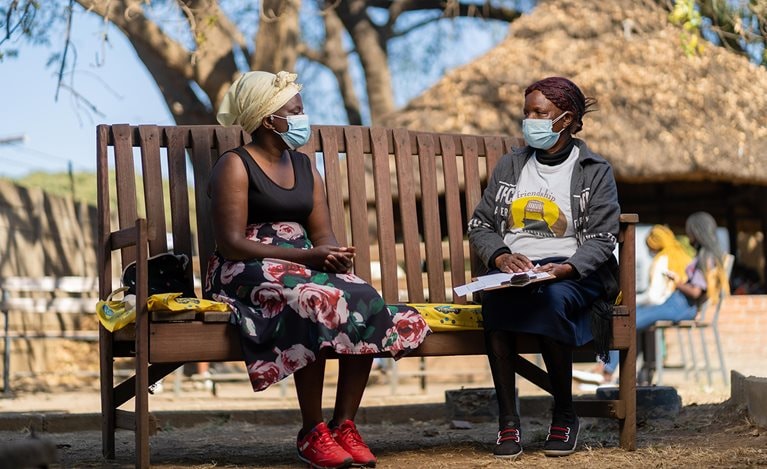 Two women wearing face masks sitting and talking on a bench in the park