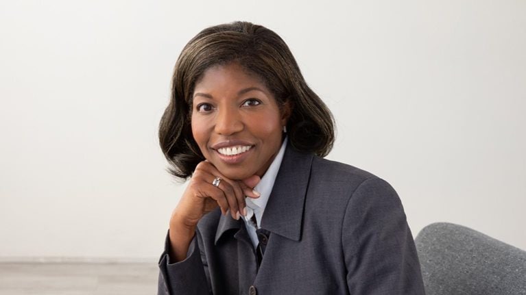 Portrait of Cheryl Pegus with a welcoming smile and seated in a office chair and her head resting on her chin.