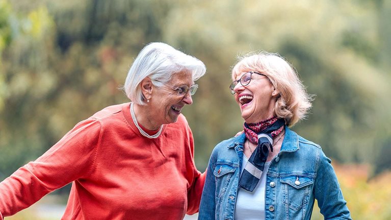 Two joyful senior women enjoy a leisurely walk in a lush park, one friend casually pushing a bicycle, both dressed in casual outdoor wear.
