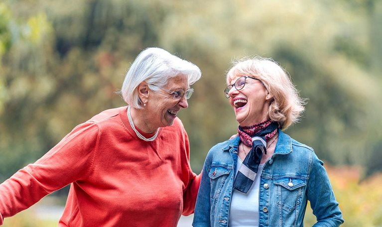 Two joyful senior women enjoy a leisurely walk in a lush park, one friend casually pushing a bicycle, both dressed in casual outdoor wear.