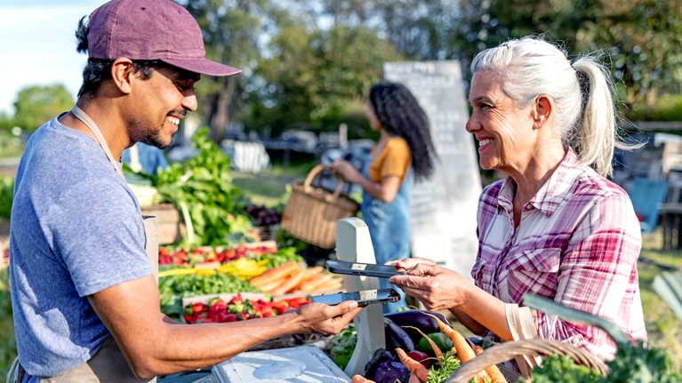 A vendor and a customer exchange a payment device across a produce stand filled with carrots, strawberries, and leafy greens at an outdoor market; another shopper stands in the background holding a basket.