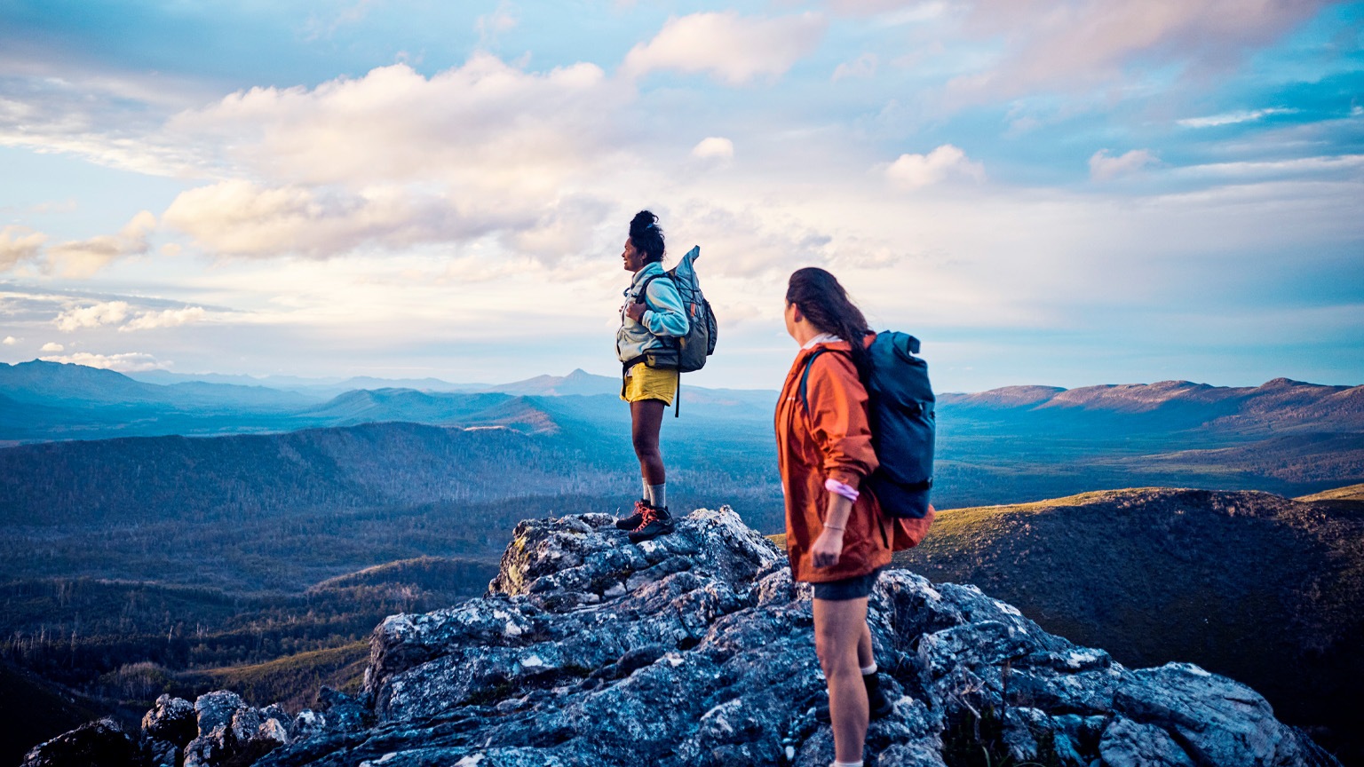 Two hikers stand on a rocky mountain peak, wearing backpacks and looking out over a vast, scenic landscape. The expansive view and soft light convey a sense of adventure, achievement, and connection with nature.