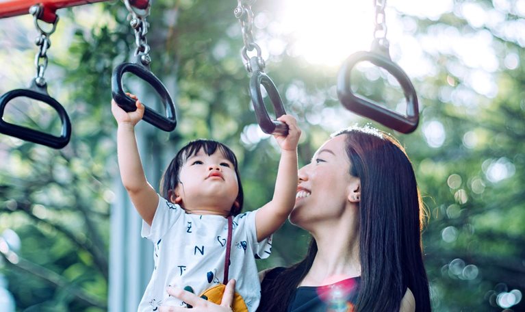 A mother smiles as she holds her young child up while the child grasps a monkey bar in an outdoor playground.