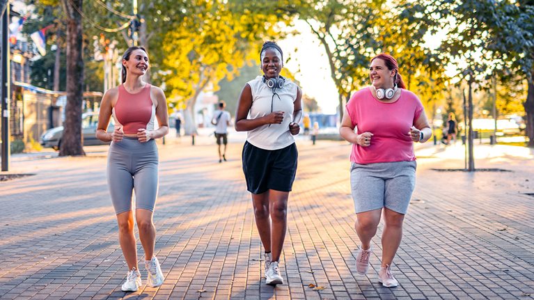 Three women with different body types are jogging together down a paved walkway, enjoying a sunny outdoor workout. They appear happy and are chatting while maintaining a comfortable pace.