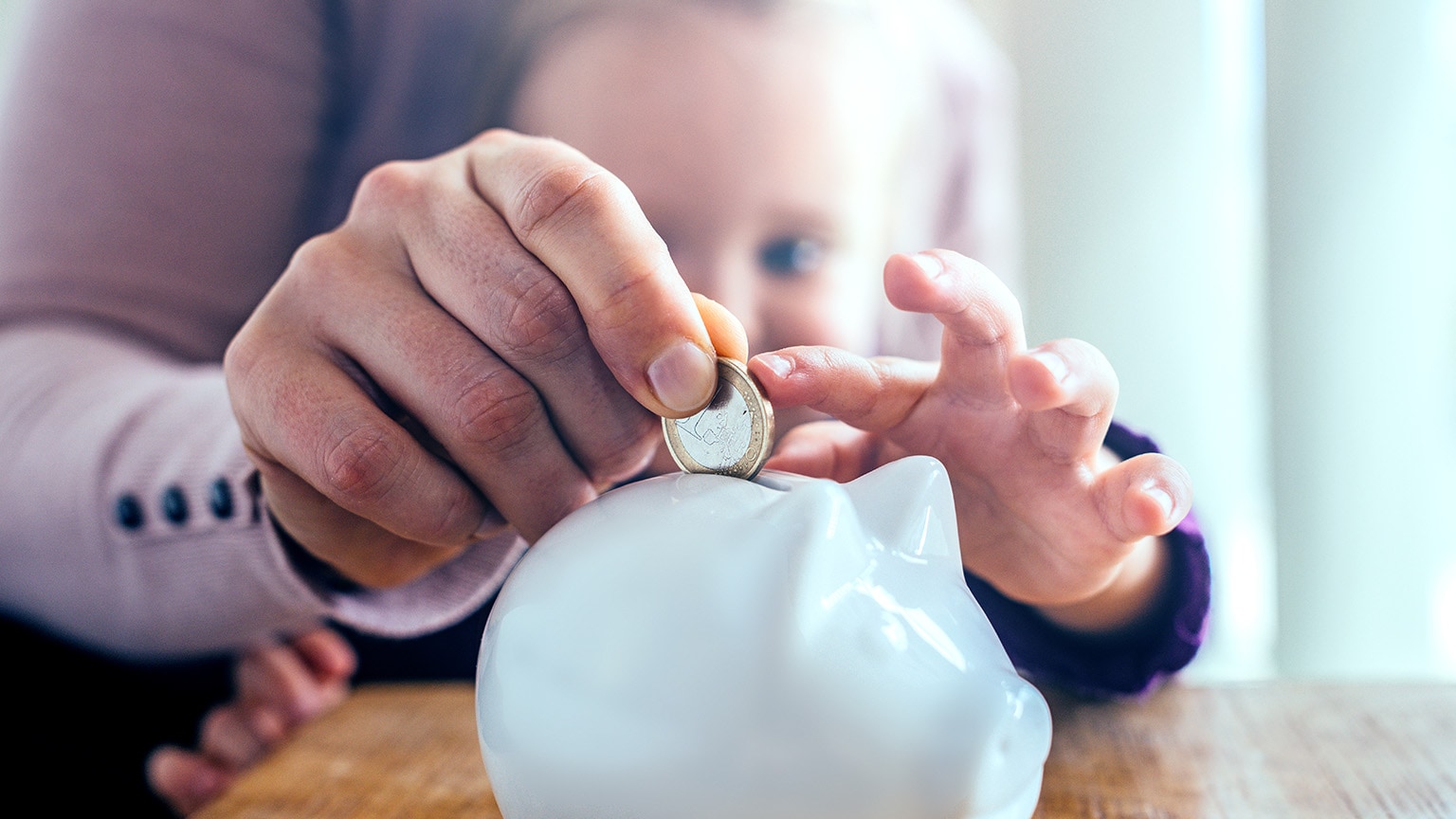 Image of a kid’s and adult’s hands putting a coin into a piggy bank.