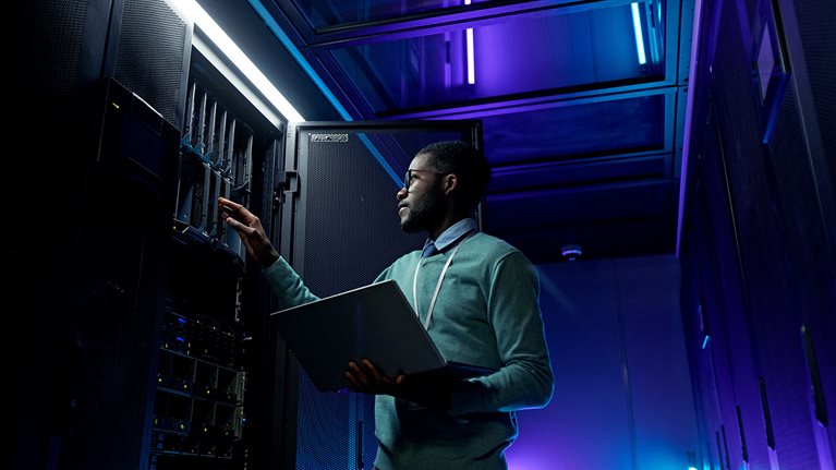 Low angle portrait of young African American data engineer working with supercomputer in server room lit by blue light and holding laptop
