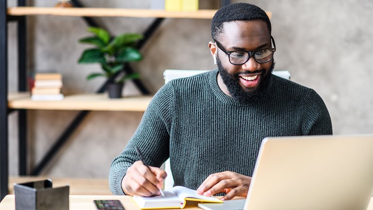 A man working on a laptop and writing notes in a book